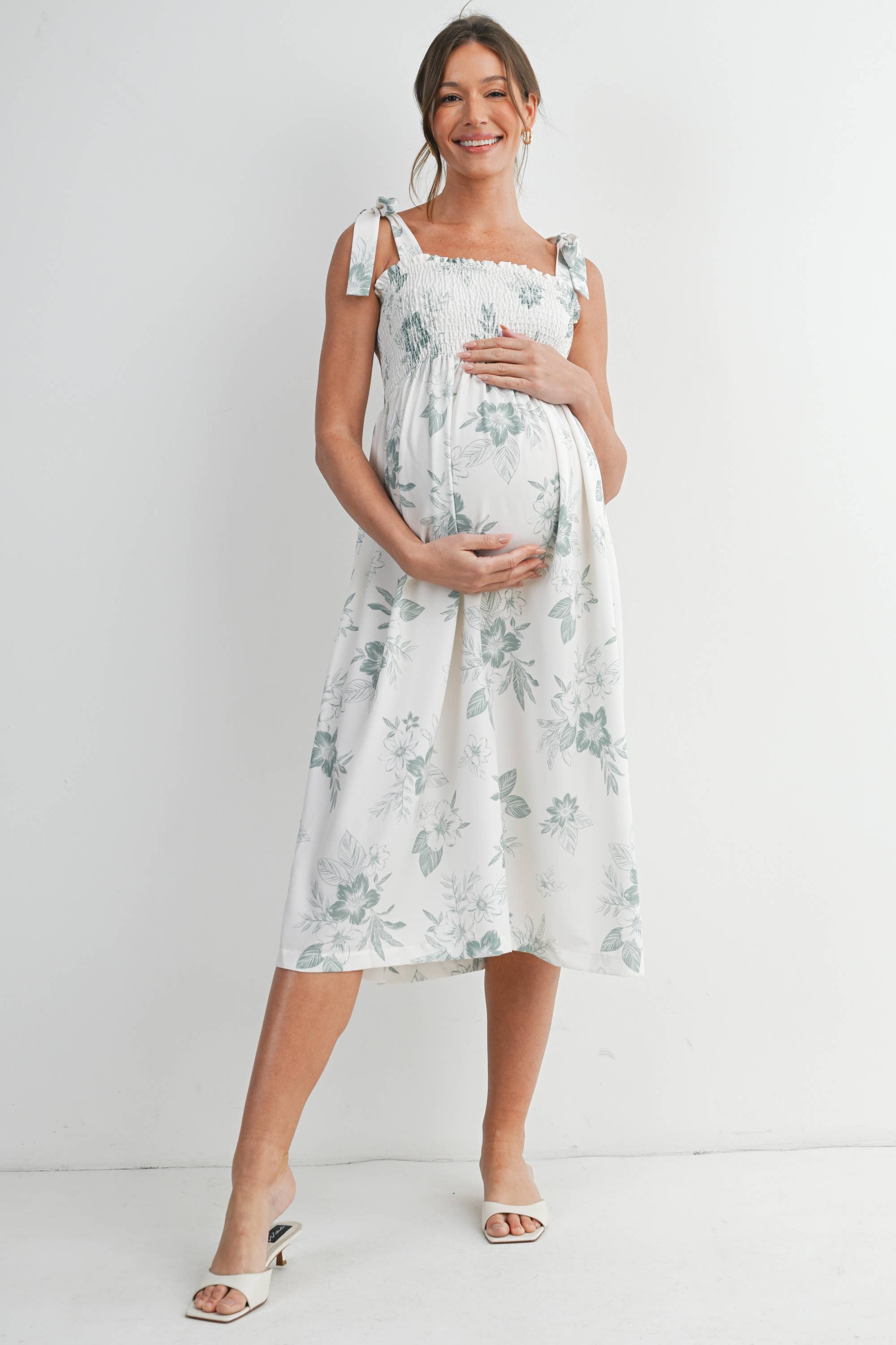 A woman standing against a white background, wearing a smocked maternity dress that is white with green green flowers 