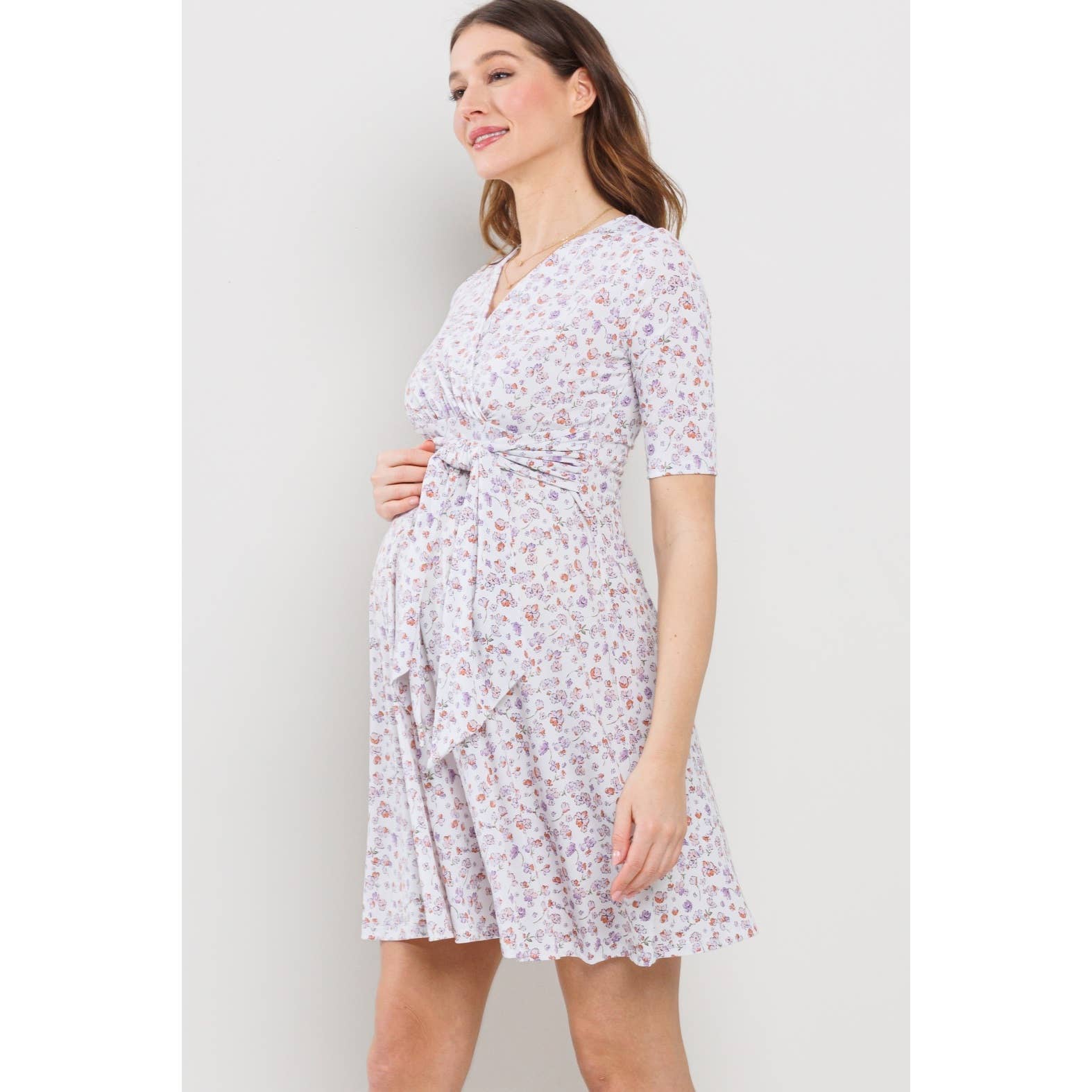 Woman facing to the left, hand on her belly wearing a floral maternity dress against a plain background