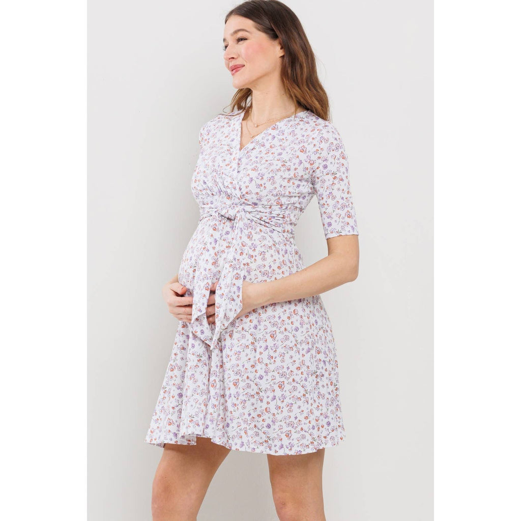 Woman facing to the right, her hands under the dress tie on her belly wearing a floral maternity dress against a plain background