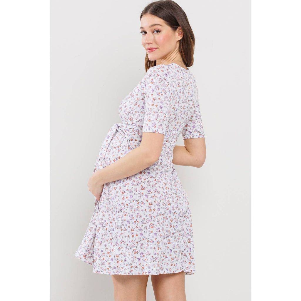Woman turned away from the camera to the left, turning her head back to face the camera wearing a maternity floral dress against a plain background