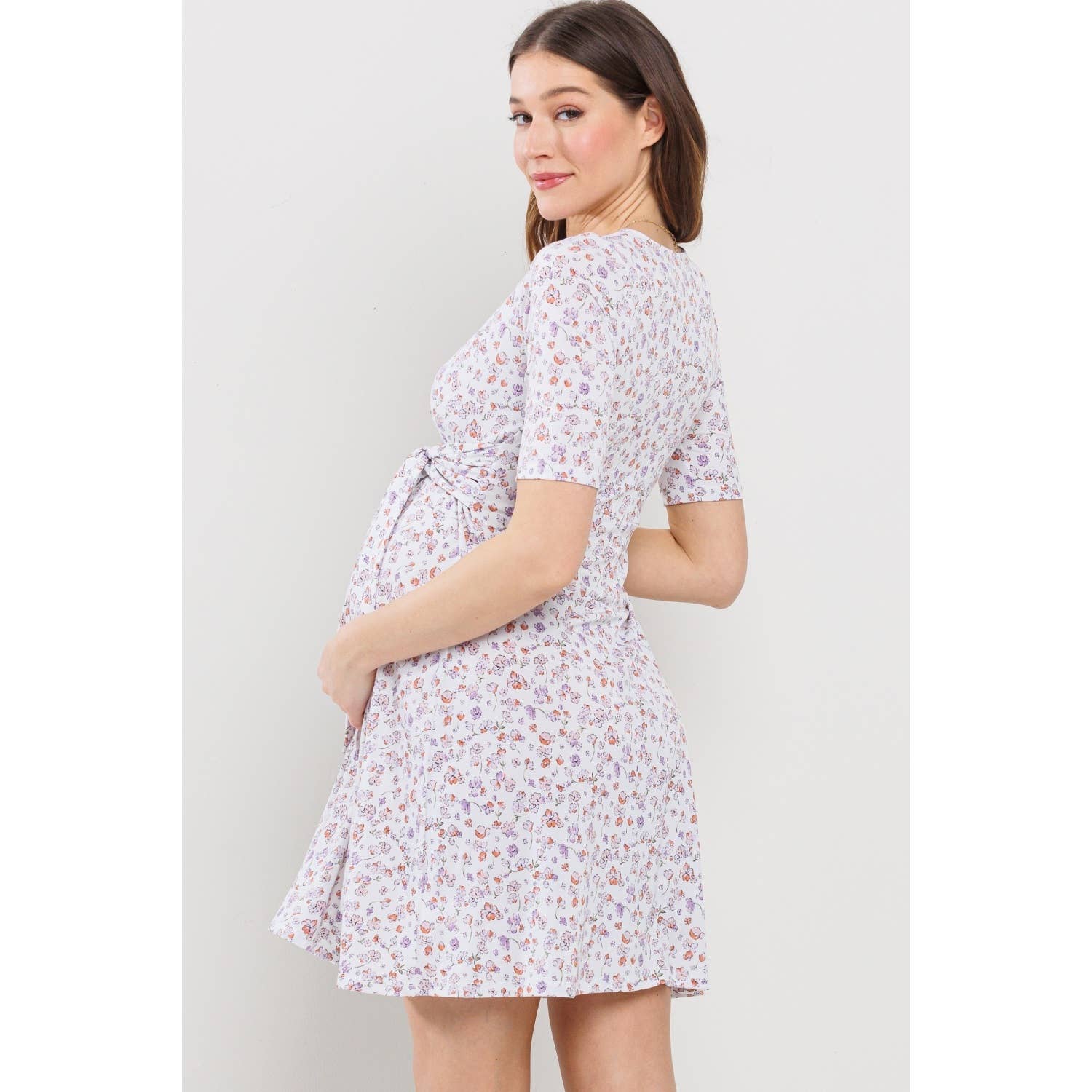 Woman turned away from the camera to the left, turning her head back to face the camera wearing a maternity floral dress against a plain background