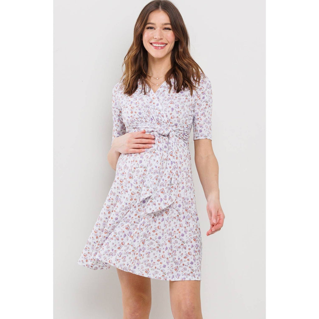 Woman with her hand on her belly wearing a white floral maternity dress against a plain background