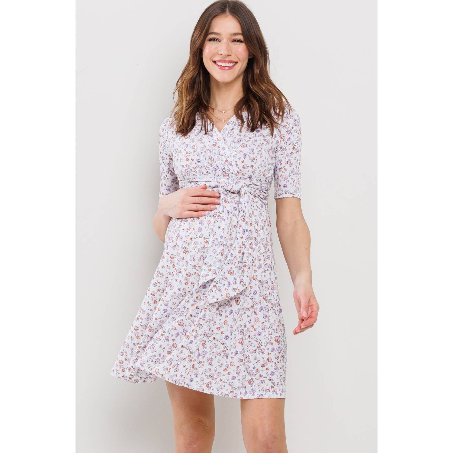 Woman with her hand on her belly wearing a white floral maternity dress against a plain background