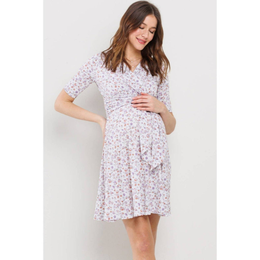 Woman standing against a gray  background, wearing a knee-length maternity dress that has pink and purple flowers