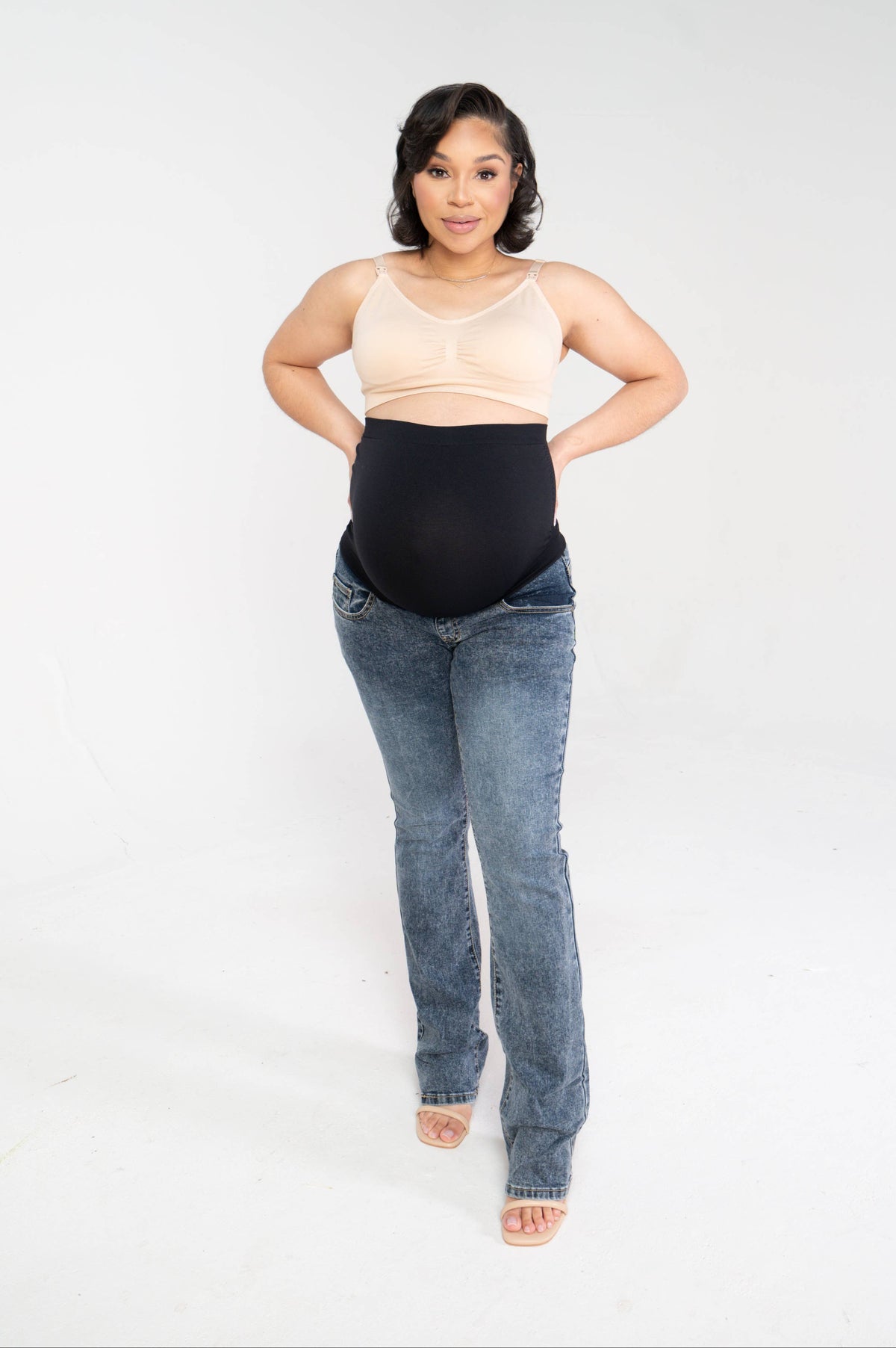 Pregnant woman standing facing the camera wearing the bootleg maternity jeans with a wide black over-the-belly panel and a beige bralette