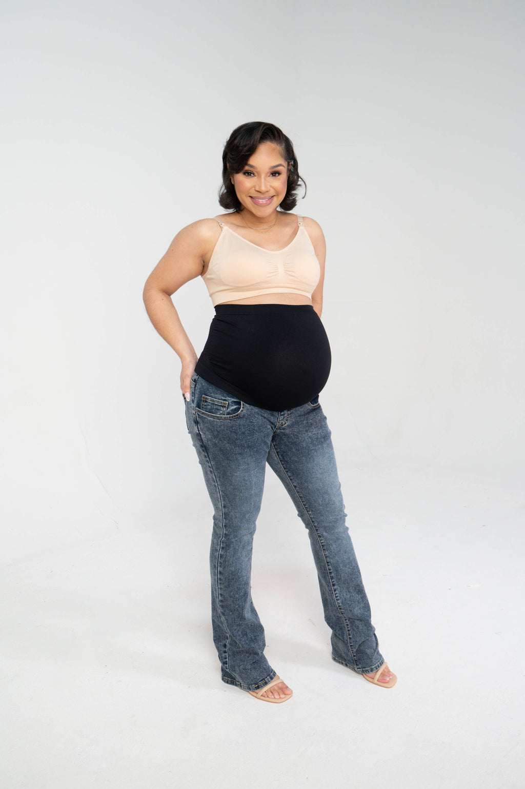 Pregnant woman standing at an angle and smiling, wearing the bootleg maternity jeans with the black over-the-belly panel