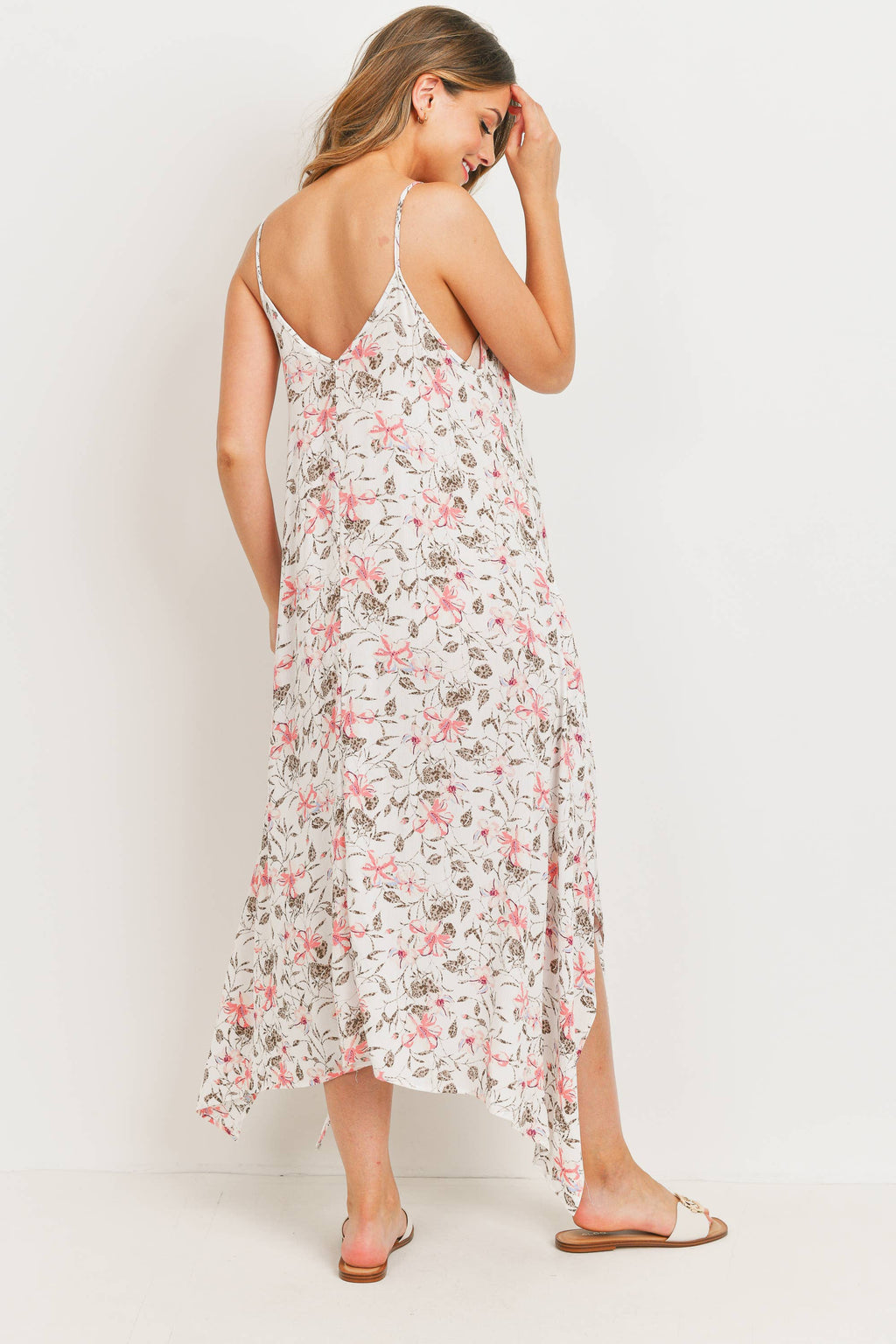 Woman wearing a tropical pink floral dress against a white background, she is faced away from the camera with her head titled down to the right, her left hand is going towards her bangs, with her right foot sticking out a little bit