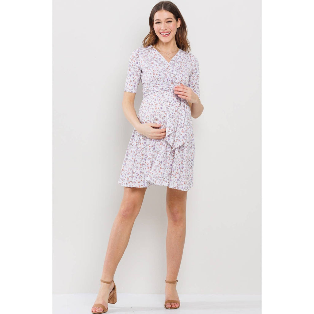 Woman wearing a floral maternity-nursing dress against a white background, she is facing the camera, and her right hand is on the top of pregnant belly and her left hand is on the bottom, she is posed with her left leg sticking out a bit while her right leg is straight 