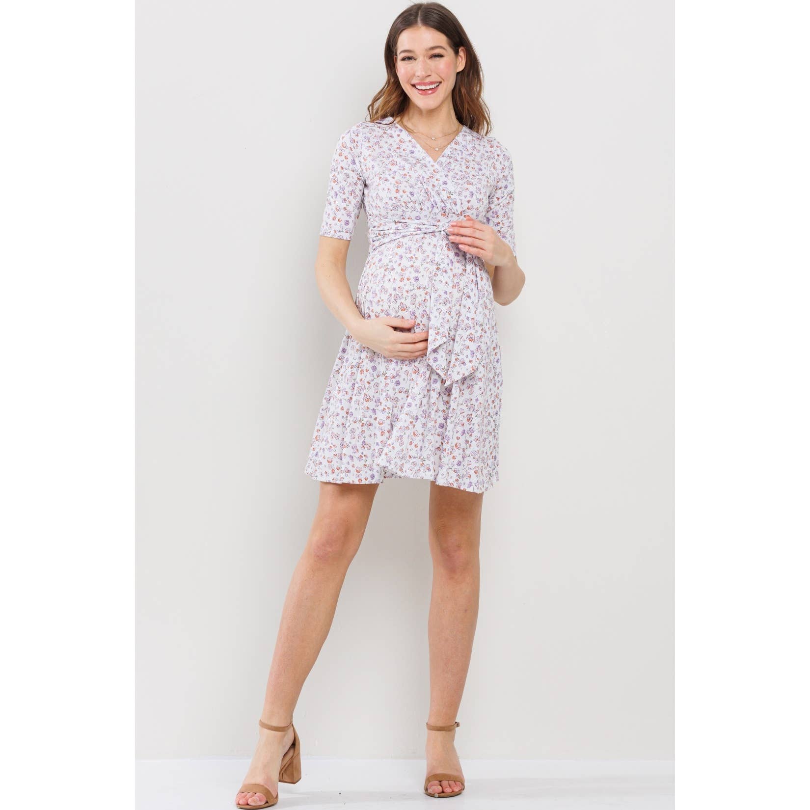 Woman wearing a floral maternity-nursing dress against a white background, she is facing the camera, and her right hand is on the top of pregnant belly and her left hand is on the bottom, she is posed with her left leg sticking out a bit while her right leg is straight 