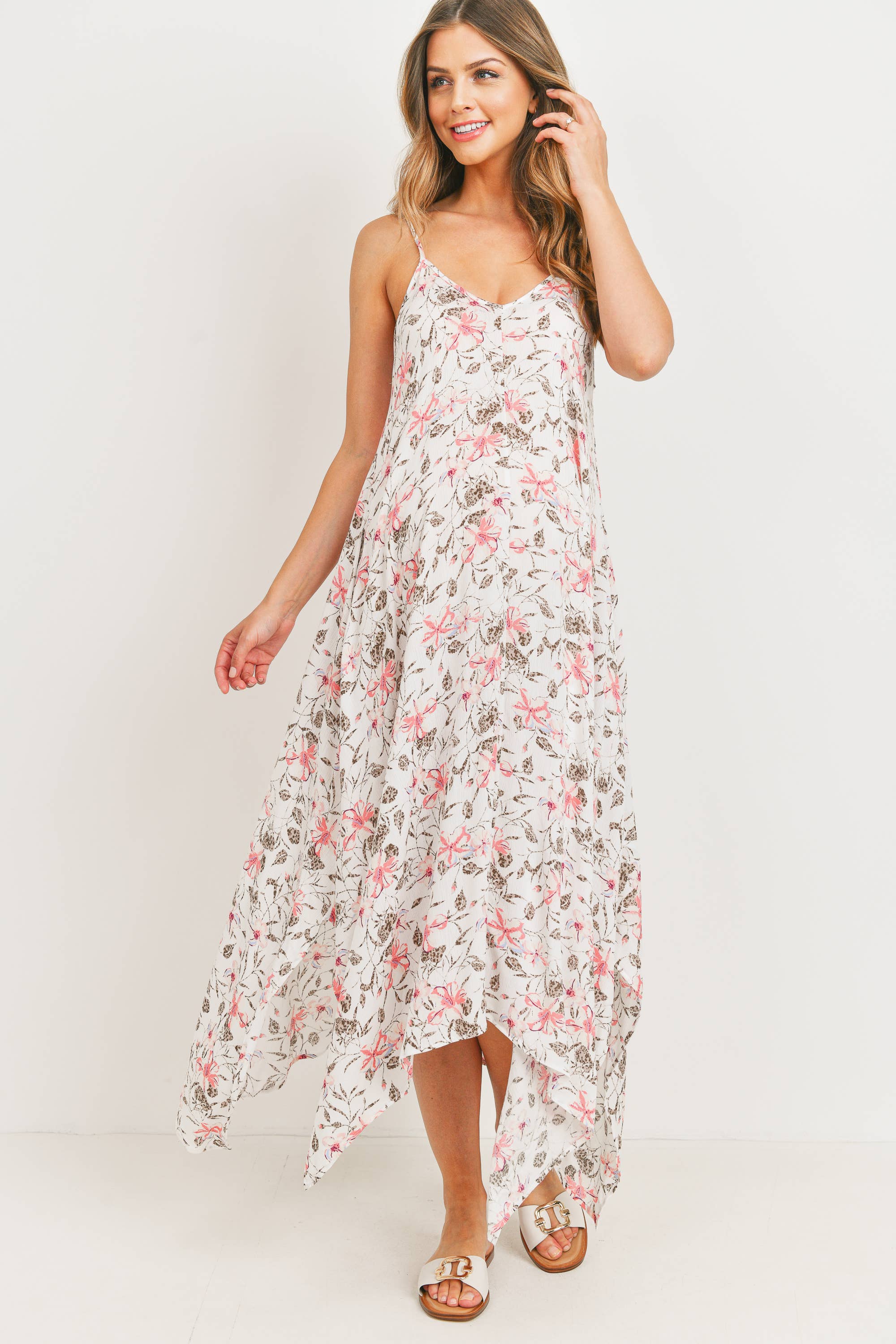 Woman wearing a tropical floral maternity dress against a white background, her right hand is in her brown/blonde hair, while her left hand is facing out to the left a little bit. Her right foot is arched back while her left leg is straight