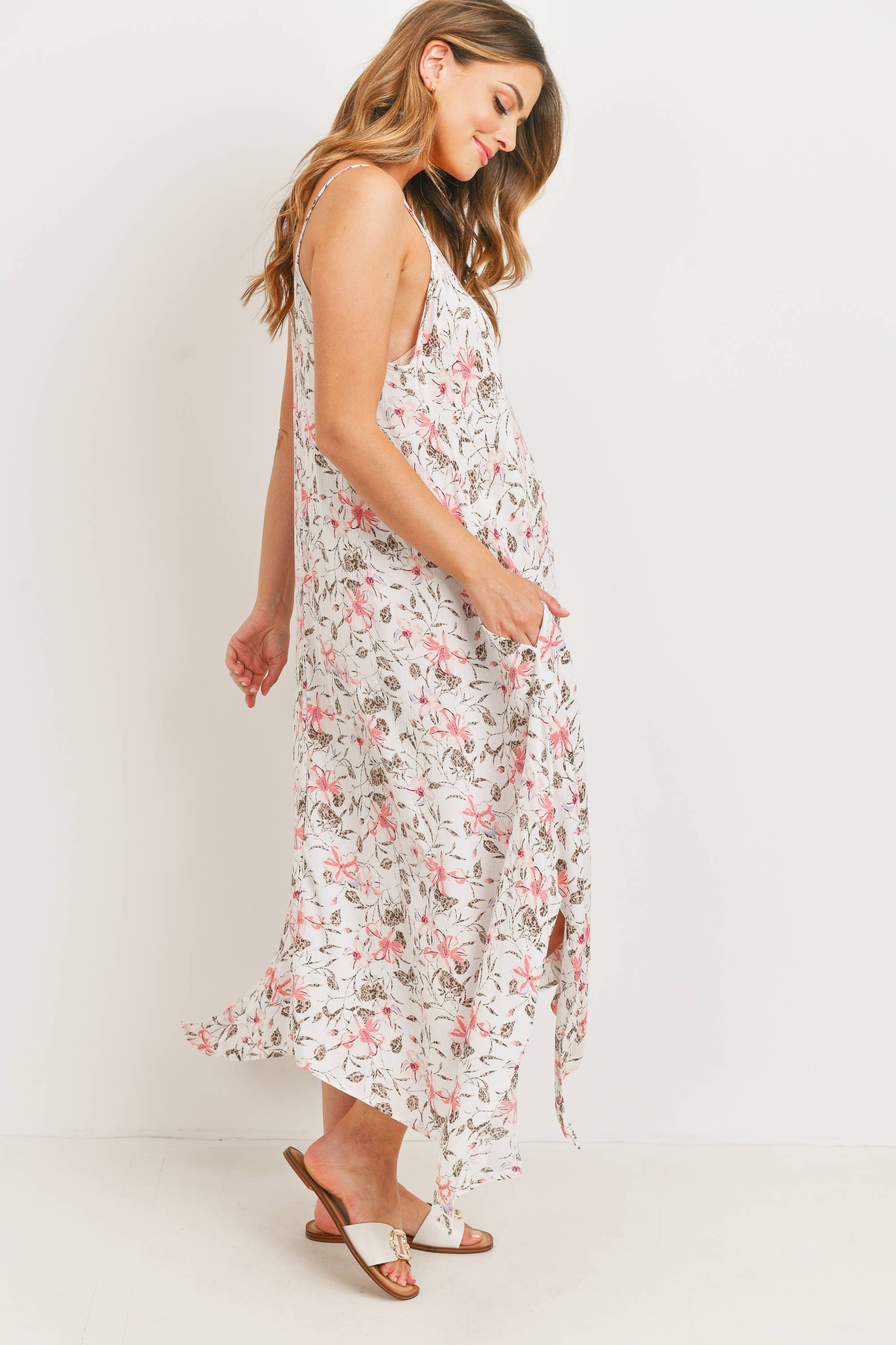 Woman facing to the right, wearing a floral dress against a white background. Her brown hair is cascading down on the right side of her face, her left hand has the dress in her hands like a Disney princess, and her right hand is tilted towards her back, her left foot is striking an arched pose