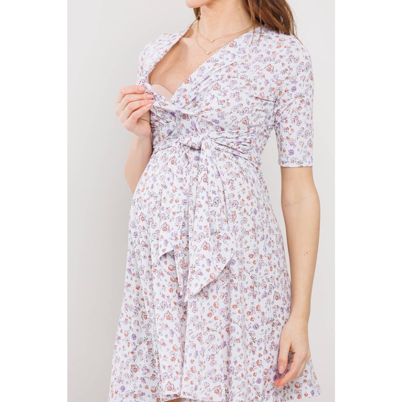 Woman wearing a floral dress against a plain background, her face is out of frame, and she is lifting down the left side of the top part on the dress to showcase that the dress is perfect for breastfeeding